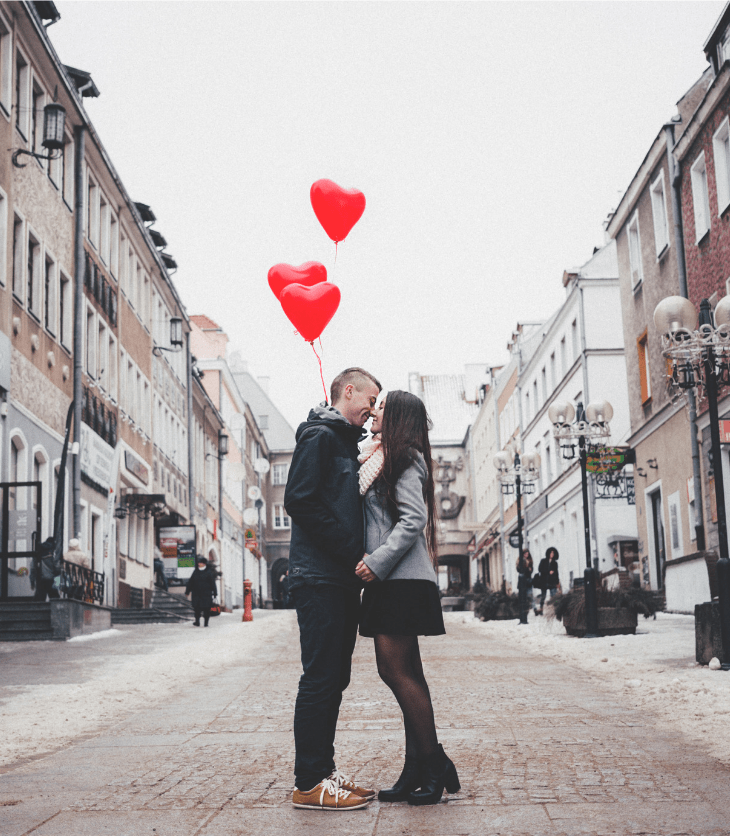couple walking in city kissing and holding red heart balloons