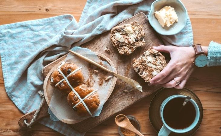 Easter hot cross buns on a plate and wooden cutting board with butter and someone holding one and a cup of coffee nearby